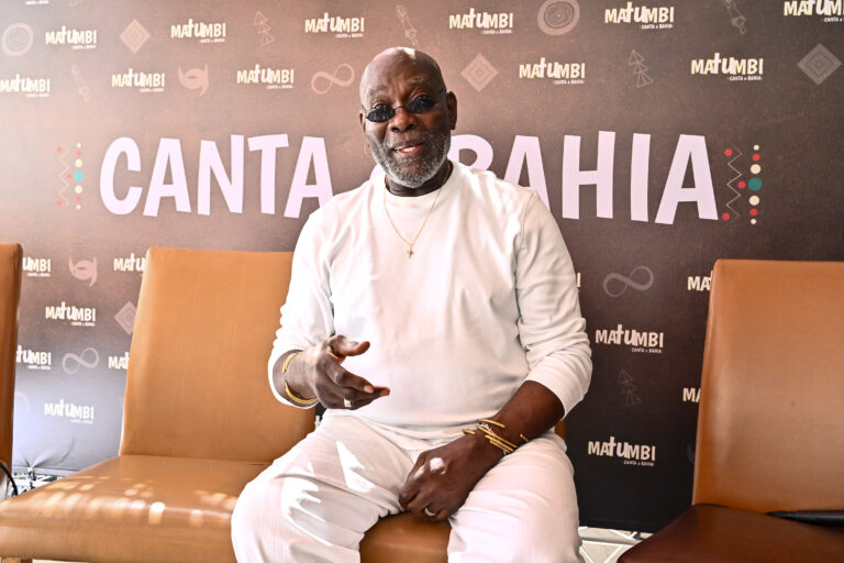 Foto do cantor Lazzo Matumbi sorrindo, sentado de roupa branca.